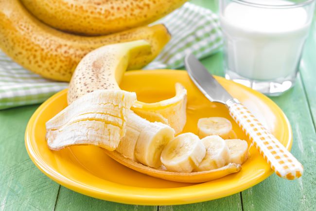 Přezrálý banán na žlutém talíři, polovina z něj je nakrájená. Banány v pozadí, na zeleném stole.