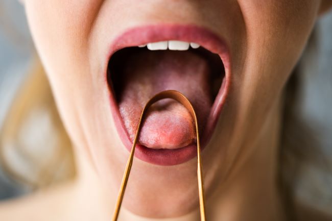 Ženská ústní dutina s vyplazeným jazykem a na něm je položena měděná škrabka na jazyk.
