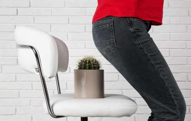 Žena v černých džínách a červeném tričku si sedá na bílou židli, na které je položen kaktus. Detailní záběr. V pozadí jsou bílé kachličky. Téma hemoroidy.