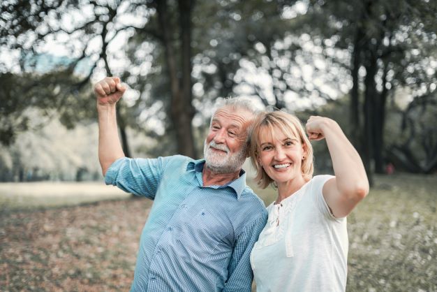 starší muž v modré košili stojí se ženou v bílém tričku v přírodě, ukazují, že jsou silní a zdraví napnutím bicepsu a usmívají se.