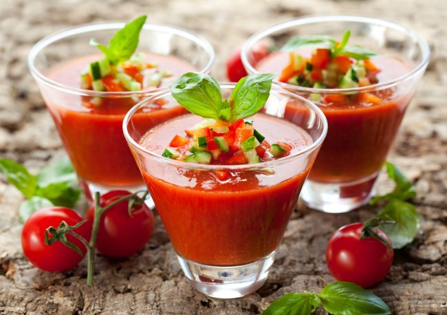 Gazpacho nápoj, z rajčat je naservírován ve skleněných sklenicích s čerstvou bazalkou na dřevěném stole, kolem jsou pokládané rajčata.