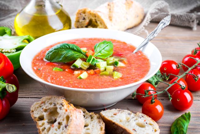 Gazpacho polévka z rajčat v bílém keramickém talíři. Na vrchu je pokrájená okurka a čerstvá bazalka. Kolem jsou čerstvá rajčata, olivový olej, chléb.