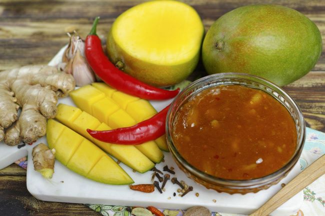 Mango chutney, čerstvé žluté mango překrojené na půlky, červené chilli jsou položeny na bílém podnosu.