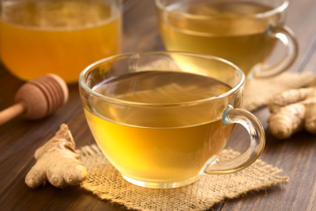 Ve skleněném šálku je žlutý zázvorový čaj, vedle šálku je položen zázvorový kořen. v pozadí je ve skleněné láhvi med.