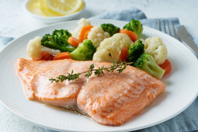 FODMAP jídlo. Na bílém talíři je naservírována dušená zelenina na páře, brokolice, květák a mrkev. Steak z lososa růžové barvy je ozdoben větvičkou tymiánu.