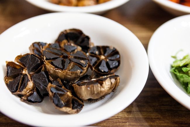 Fermentovaný černý česnek na bílém talíři jako součást zdravé výživy. Fermentovaný černý česnek má blahodárné účinky na zdraví