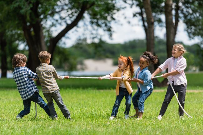 Dva kluci a tři dívky stojí na trávníku v přírodě, tráva je krásně zelená, hrají si s lanem dětskou hru.