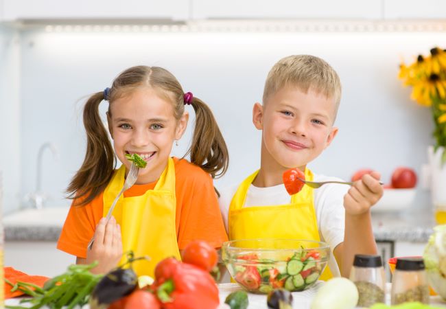 Blonďatý chlapeček a hnědovlasá holčička se dvěma copy sedí za stolem, kde je hodně zeleniny, oba se usmívají a jedí zeleninový salát.