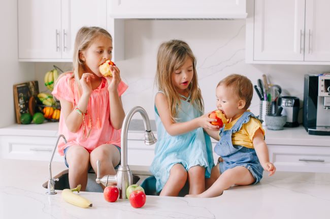 Tři malé děti sedí na kuchyňské lince. Blondýna kouše jablko, hnědovláska krmí nejmladší dítě broskví.